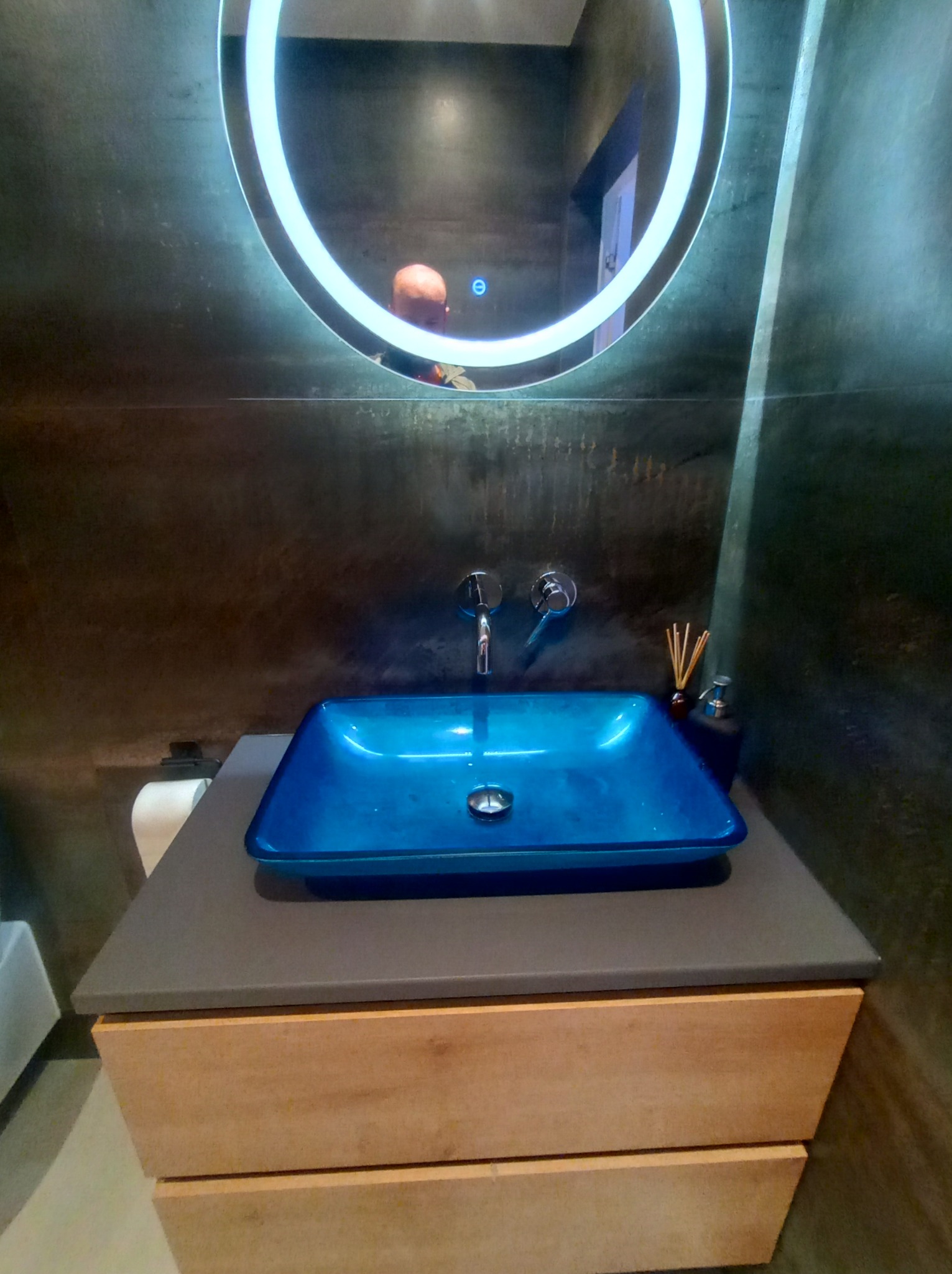 Niebieska, szklana umywalka nablatowa w nowoczesnej łazience w Myślenicach, z podświetlanym lustrem i szarą ścianą o fakturze betonu.