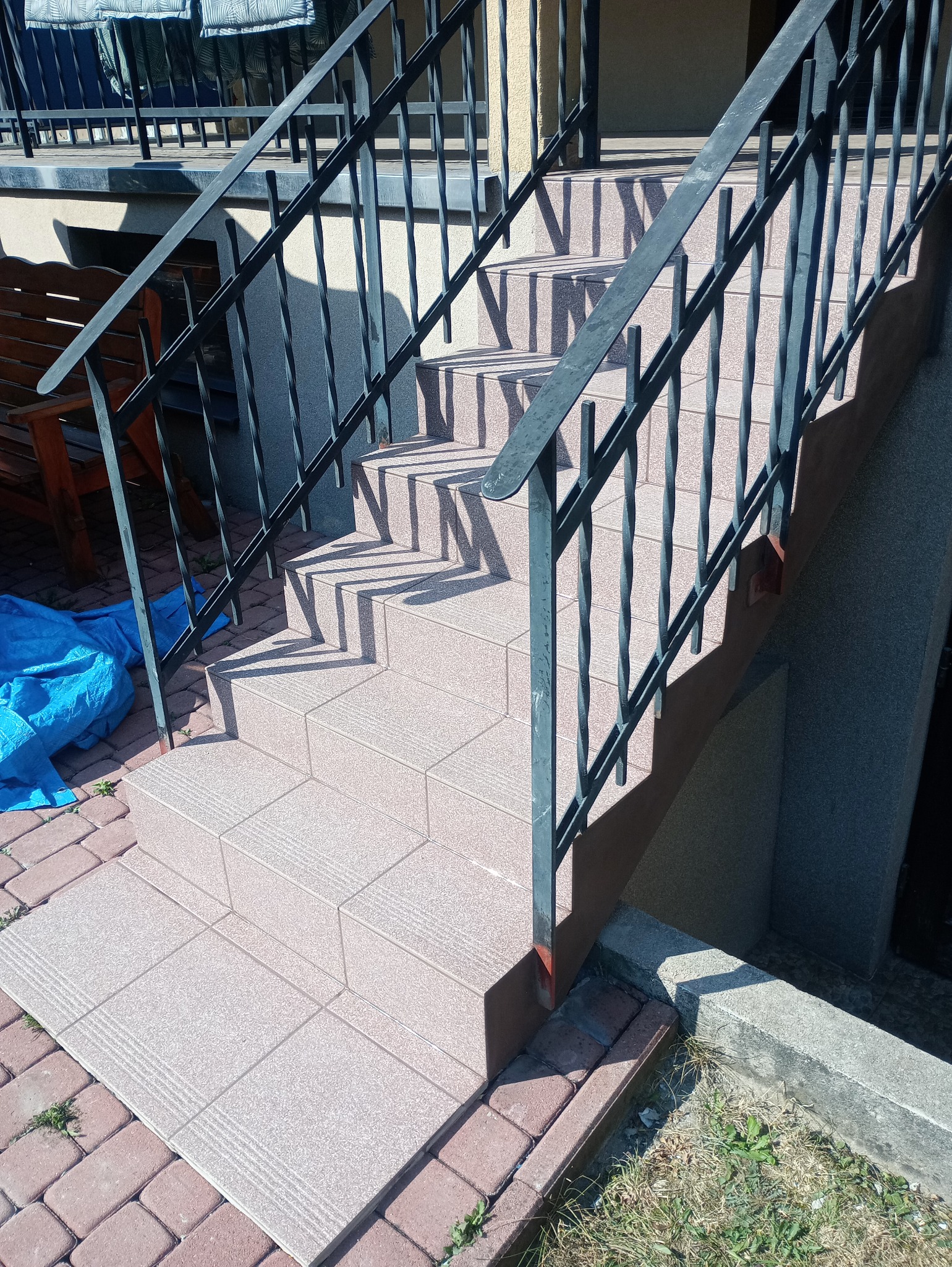 Zewnętrzne schody wyłożone jasnymi, kwadratowymi płytkami z delikatnym wzorem, z czarną metalową balustradą, rzucające cienie na stopnie; obok widoczny fragment drewnianej ławki i niebieska plandeka.