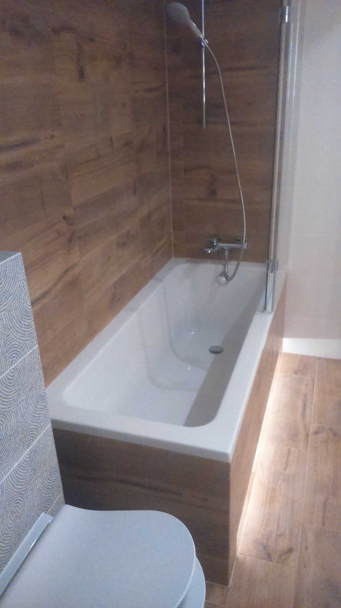 Biała wanna obudowana panelami imitującymi drewno, z zamontowanym szklanym parawanem i baterią, obok toaleta z płytkami w geometryczny wzór, całość w łazience wyłożonej płytkami imitującymi drewno.