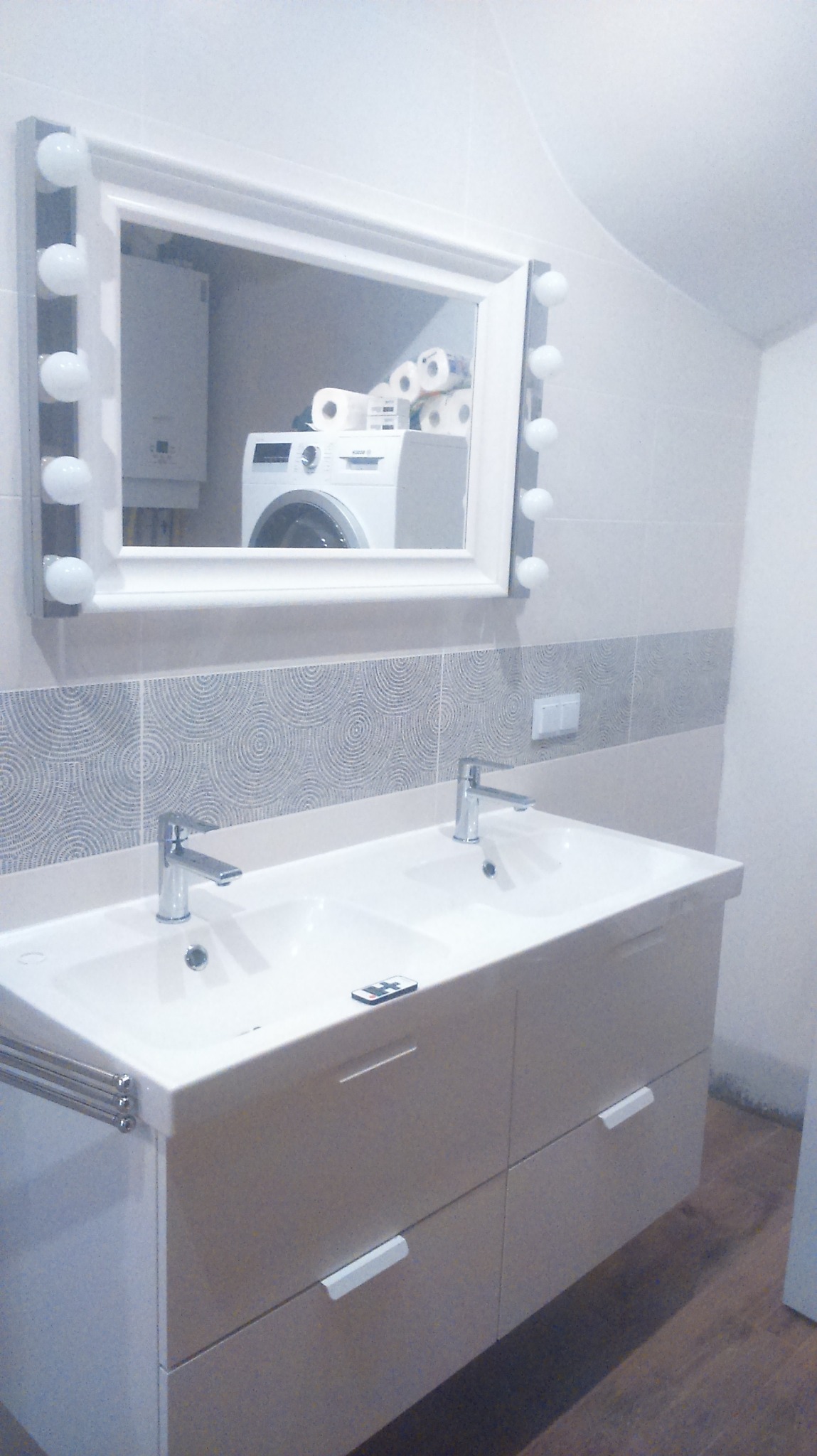 Nowoczesna łazienka z podwójną umywalką na szafce z szufladami, lustro z oświetleniem w stylu hollywoodzkim odbija pralkę i zapas papieru toaletowego.