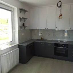 Nowoczesna kuchnia z szarymi dolnymi szafkami i białymi górnymi, złota bateria umywalkowa, marmurowe panele ścienne, drewniana podłoga i minimalistyczna lampa wisząca z trzema żarówkami.