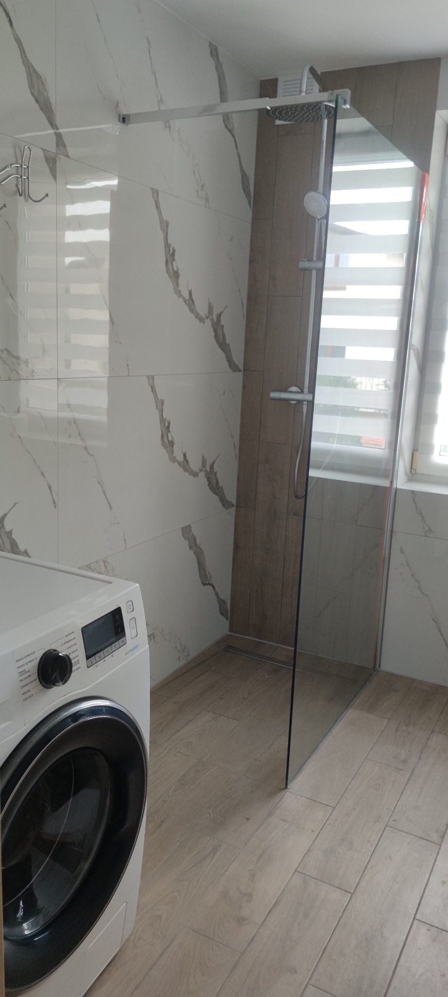 Nowoczesna łazienka z kabiną prysznicową typu walk-in, wykończona płytkami imitującymi marmur i drewnem, z widoczną pralką automatyczną.