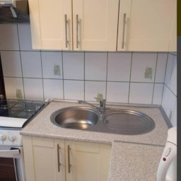 Weronika Szecówka - Kuchenny zlew dwukomorowy ze stali nierdzewnej, bateria, płyta grzewcza i białe szafki z podłużnymi uchwytami na tle białych płytek z delikatnym zielonym wzorem.