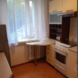 Weronika Szecówka - Kuchnia z beżowymi szafkami, brązowymi płytkami podłogowymi i blatem roboczym przy oknie z białymi zasłonami.