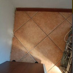 Weronika Szecówka - Ukośne ułożenie brązowych płytek podłogowych, widoczna listwa przypodłogowa, fragment szafki i pralki, nierówne wykończenie ściany.