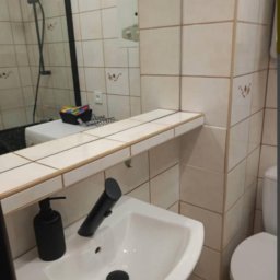 Weronika Szecówka - Umywalka z czarną armaturą i dozownikiem mydła w łazience wyłożonej beżowymi płytkami z dekoracyjnymi wzorami, widok z perspektywy lustra odbijającego prysznic.