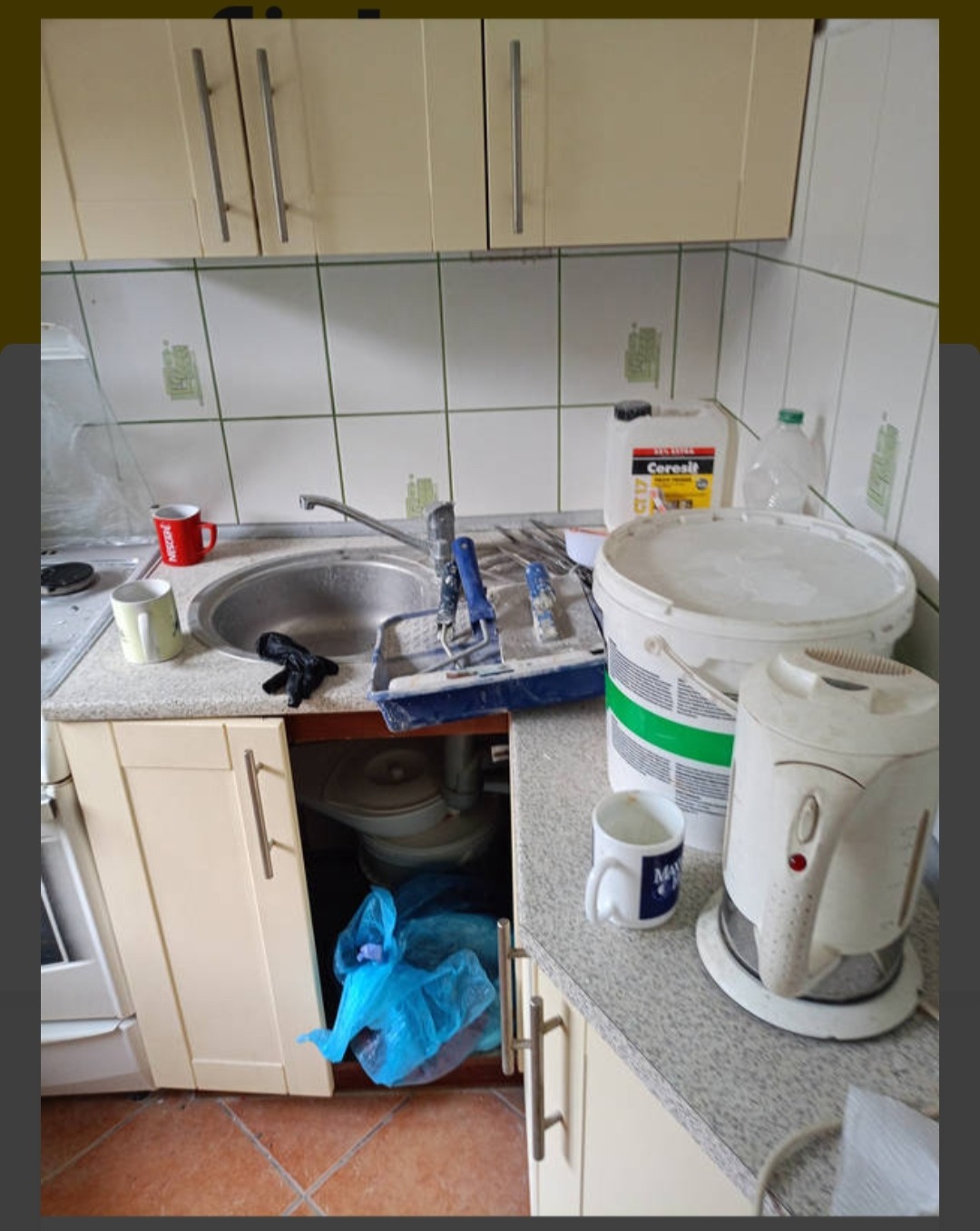 Umywalka kuchenna z narzędziami malarskimi, wiadrem farby Ceresit, kubkiem i czajnikiem elektrycznym po remoncie, widoczne zabrudzenia na płytkach i podłodze.