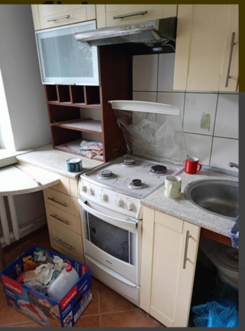 Brudna kuchnia przed sprzątaniem: widoczny zabrudzony blat, płyta gazowa z resztkami, pudełko z odpadami na podłodze, plamy na kafelkach i ogólny bałagan.