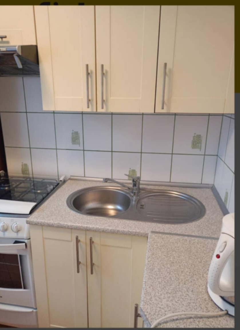 Kuchenny zlew dwukomorowy ze stali nierdzewnej, bateria, płyta grzewcza i białe szafki z podłużnymi uchwytami na tle białych płytek z delikatnym zielonym wzorem.