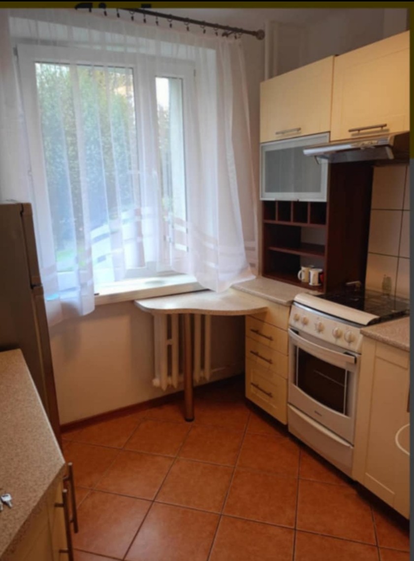 Kuchnia z beżowymi szafkami, brązowymi płytkami podłogowymi i blatem roboczym przy oknie z białymi zasłonami.