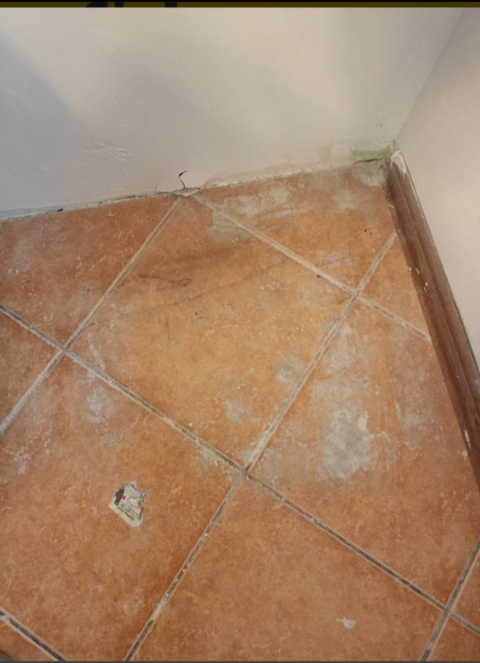 Fragment podłogi wyłożonej brązowymi, kwadratowymi płytkami ceramicznymi przed gruntownym wysprzątaniem, widoczne zabrudzenia, resztki kleju i oderwana naklejka na powierzchni płytek, ślady po...