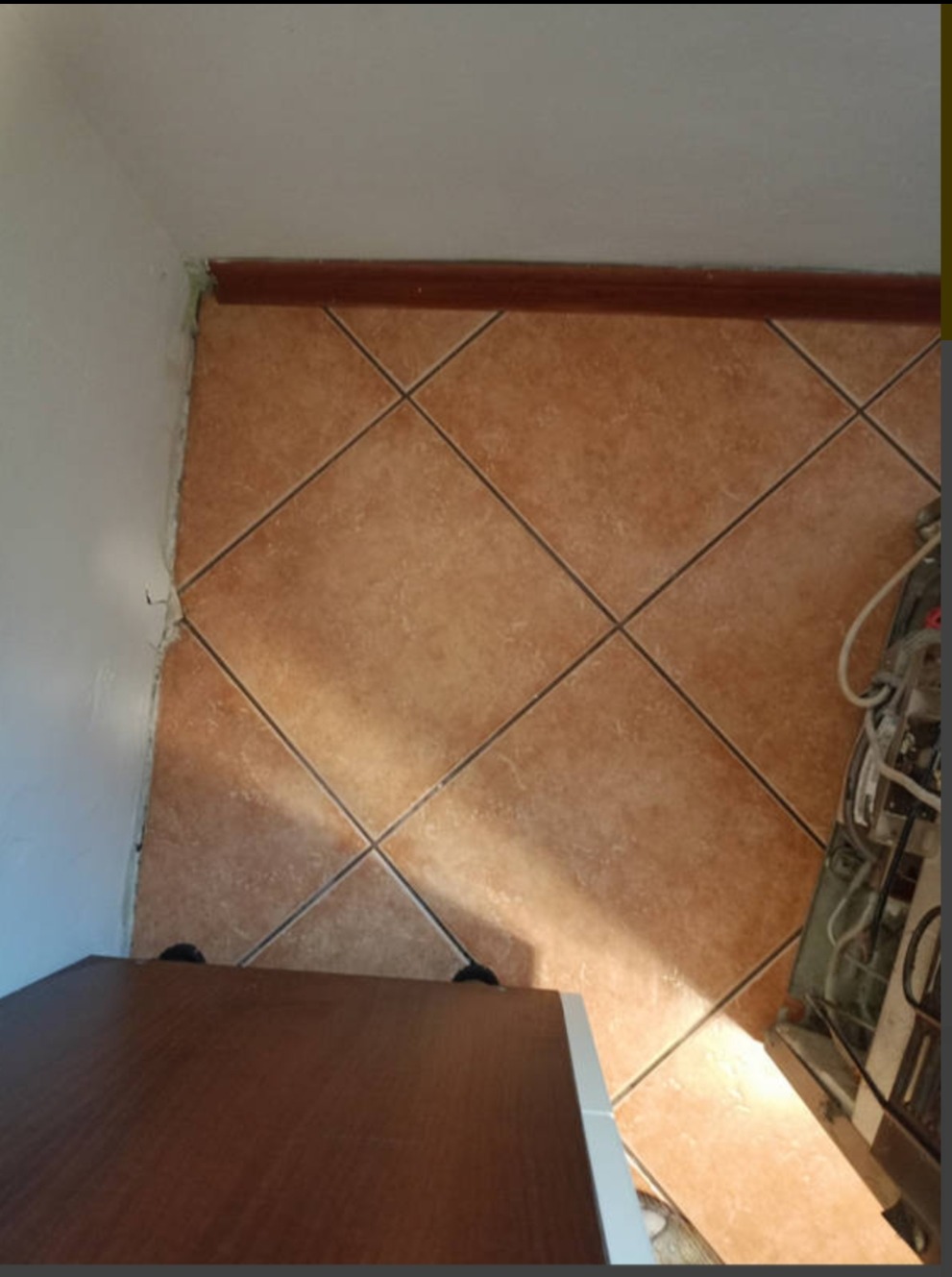 Ukośne ułożenie brązowych płytek podłogowych, widoczna listwa przypodłogowa, fragment szafki i pralki, nierówne wykończenie ściany.