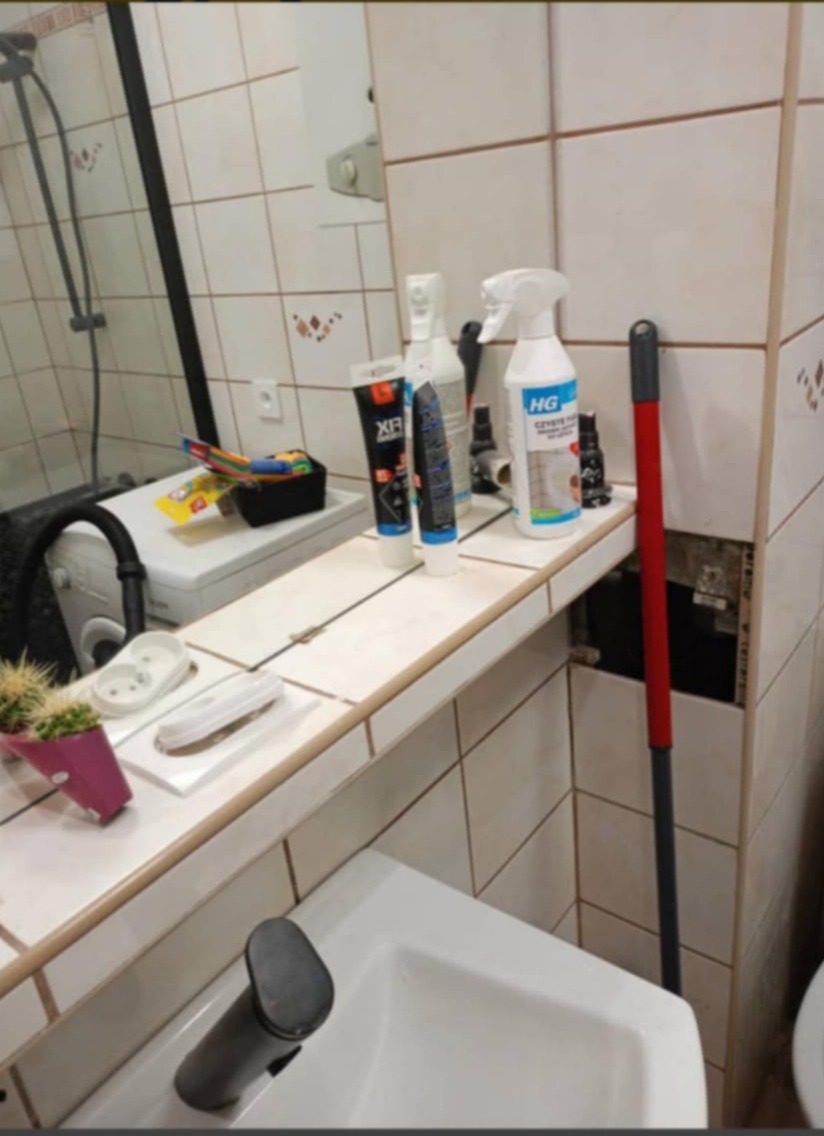 Umywalka z baterią, nad nią półka z kosmetykami, środkami czystości HG i mopem opartym o ścianę z uszkodzonymi płytkami.