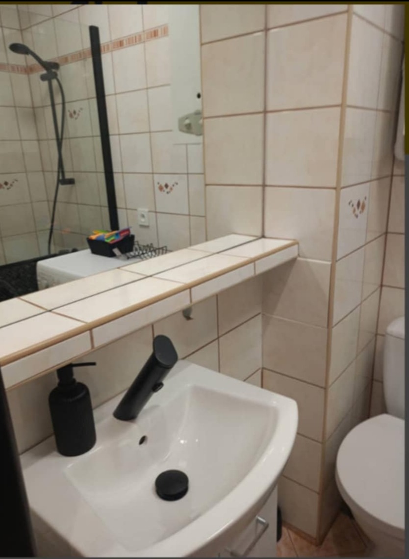 Umywalka z czarną armaturą i dozownikiem mydła w łazience wyłożonej beżowymi płytkami z dekoracyjnymi wzorami, widok z perspektywy lustra odbijającego prysznic.