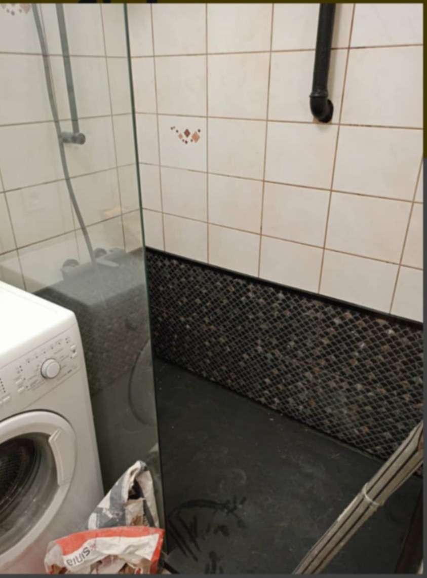 Niedokładnie umyta kabina prysznicowa z widocznymi zaciekami, obok pralka i resztki gruzu w worku, na podłodze plamy.