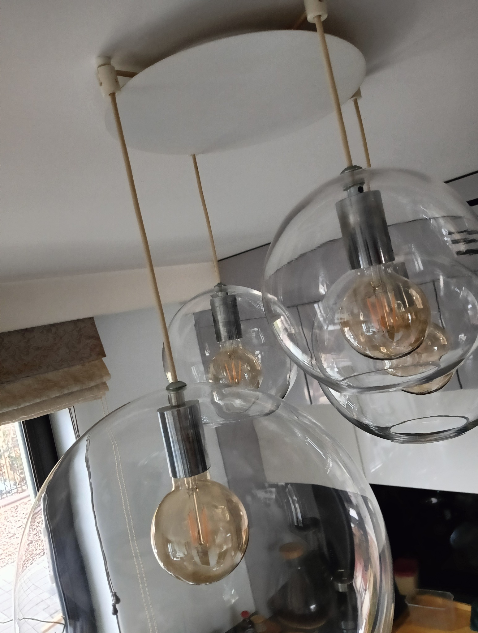 Trzy szklane lampy wiszące z żarówkami Edisona, widok z dołu, z fragmentem rolety okiennej w tle.