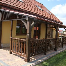 FORMA pracownia drewna - Zadaszony taras przy domu z drewnianą balustradą i kolumnami. Widoczne brązowe elementy konstrukcyjne i czerwona dachówka. Ujęcie z poziomu trawnika.