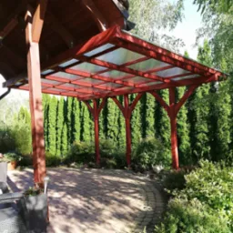 Czerwona pergola z przezroczystym dachem na brukowanym patio, otoczona zielenią. Widoczne meble ogrodowe i wysokie tuje w tle. Słoneczny dzień.