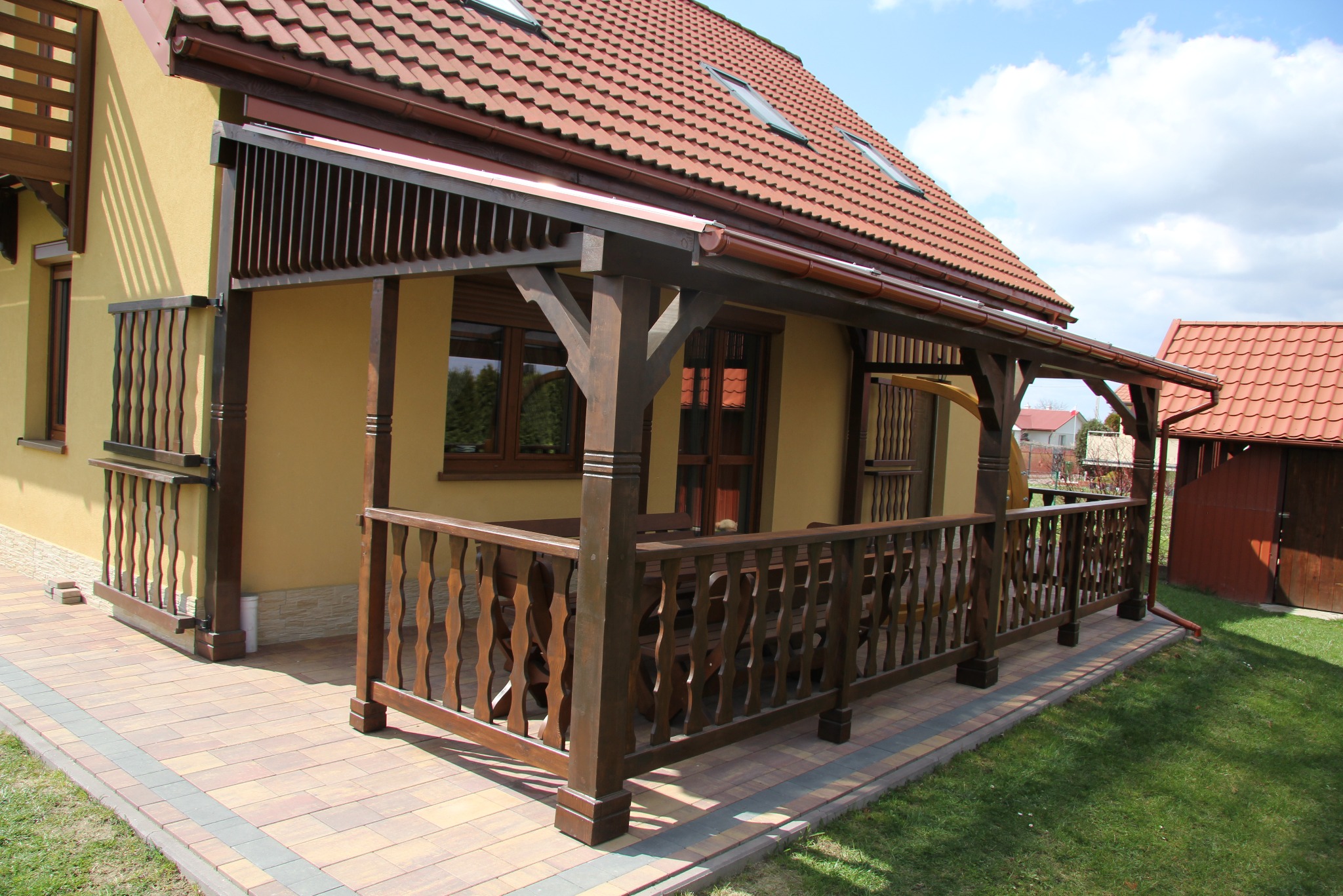 Zadaszony taras przy domu z drewnianą balustradą i kolumnami. Widoczne brązowe elementy konstrukcyjne i czerwona dachówka. Ujęcie z poziomu trawnika.