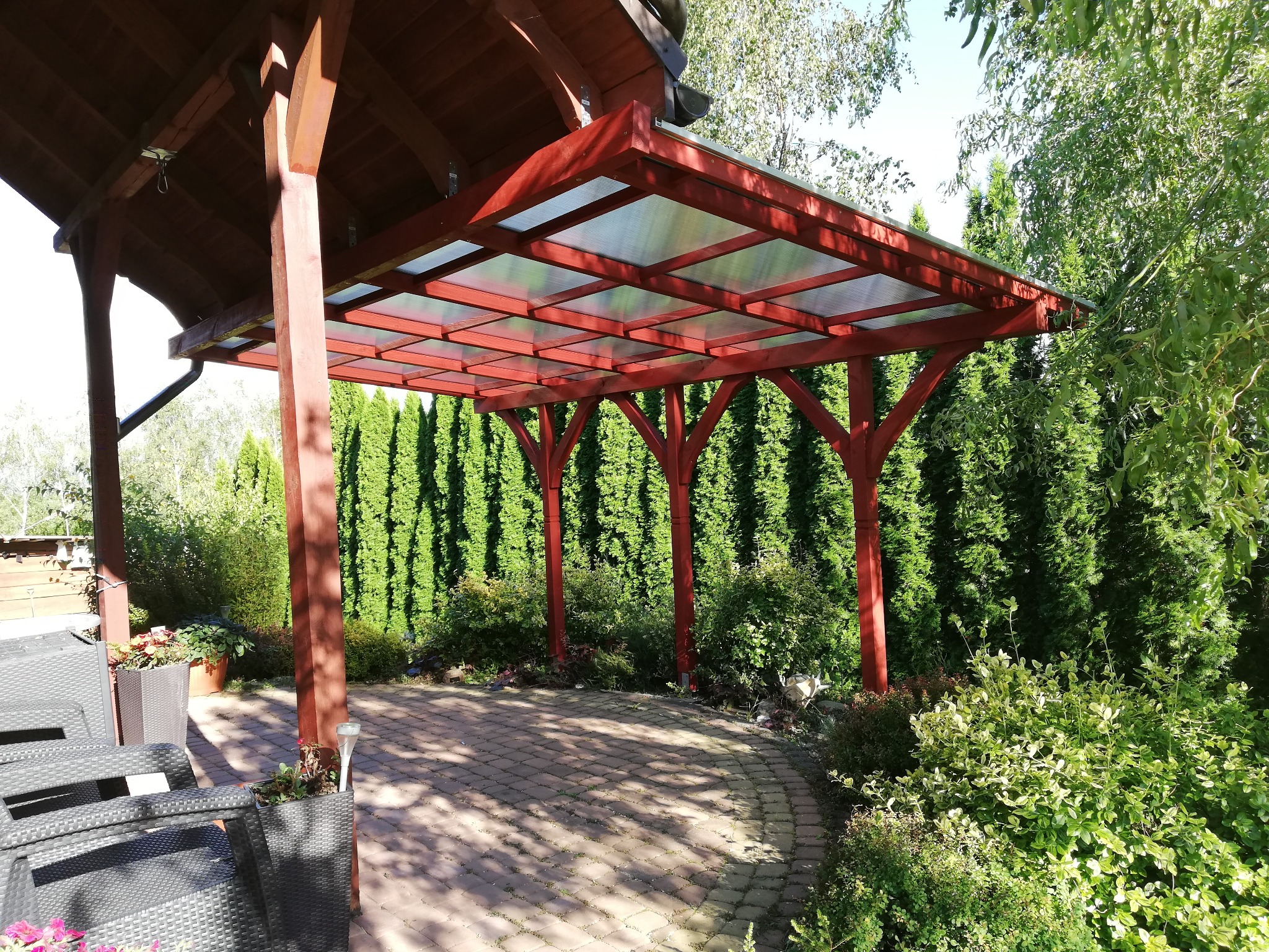 Czerwona pergola z przezroczystym dachem na brukowanym patio, otoczona zielenią. Widoczne meble ogrodowe i wysokie tuje w tle. Słoneczny dzień.