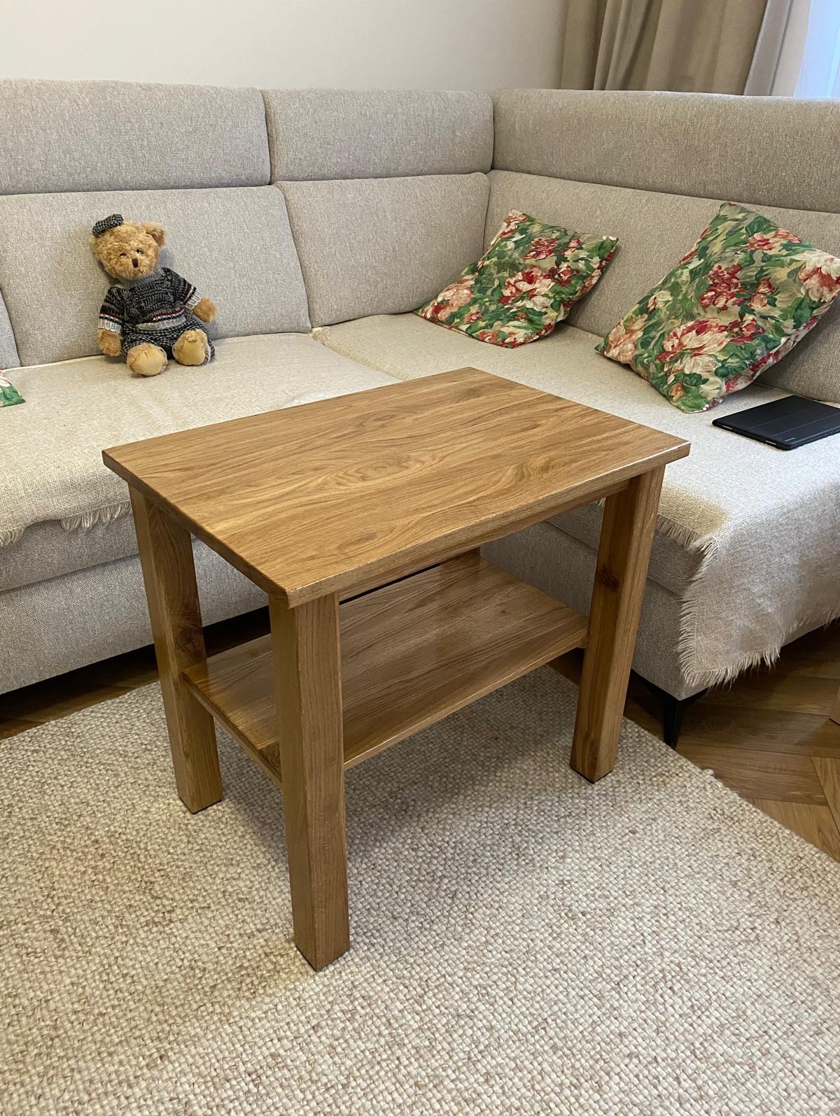 Drewniany stolik kawowy z półką, wykonany z jasnego drewna, stoi na beżowym dywanie. W tle szara sofa z poduszkami w kwiaty i pluszowy miś z kokardką.