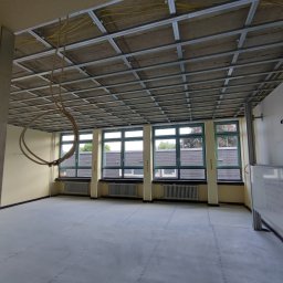 Montaris - Metalowa konstrukcja sufitowa w trakcie montażu, widoczne kable elektryczne, jasne ściany i duże okna w tle, biała tablica z notatkami po prawej stronie.