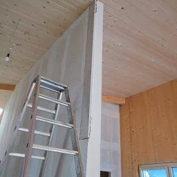 Montaris - Wnętrze w trakcie prac wykończeniowych: ściana z płyt gipsowo-kartonowych, drewniany sufit, aluminiowa drabina oparta o ścianę, widoczne instalacje elektryczne w ścianie, fragment okna balkonowego...