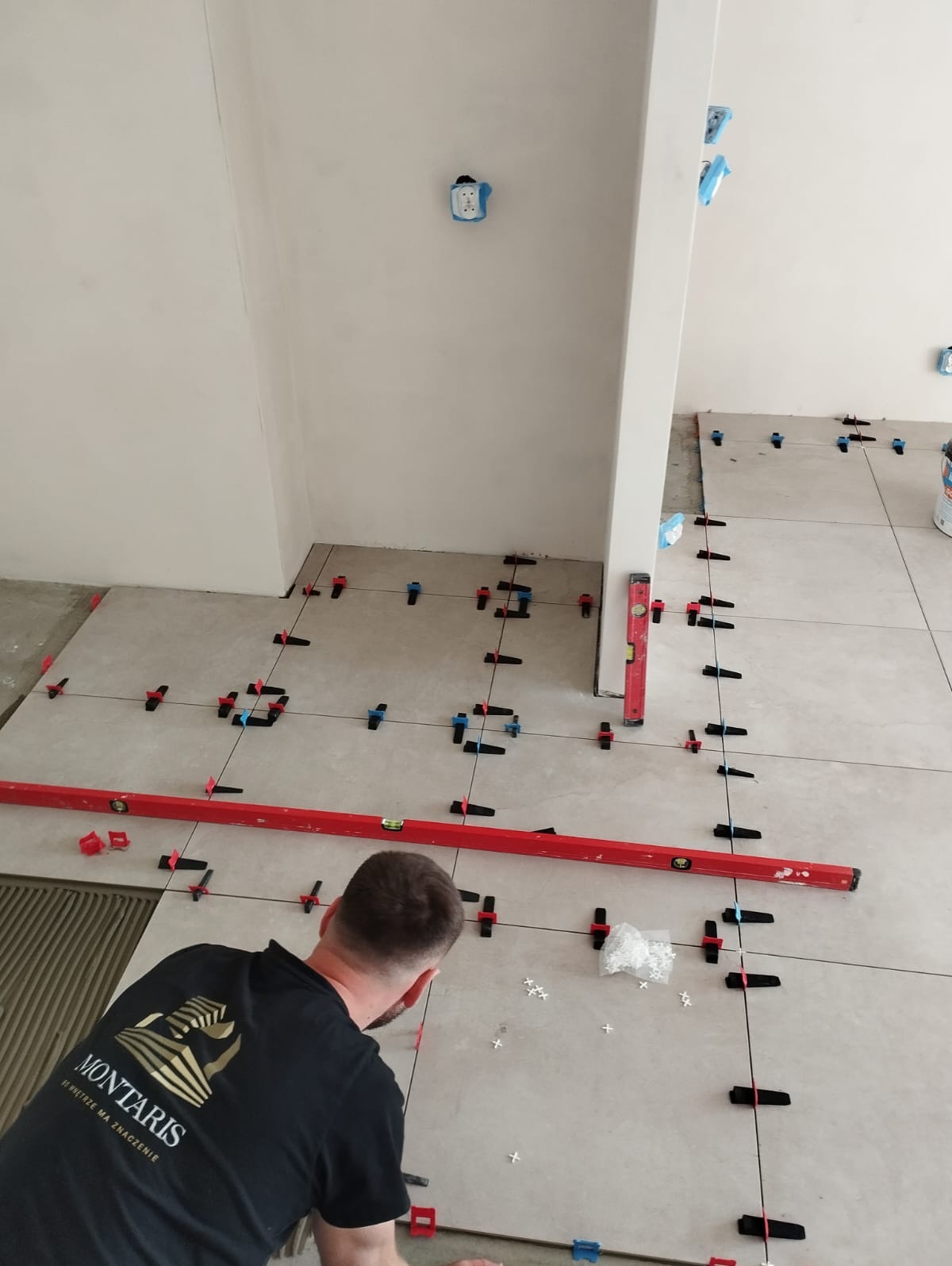 Układanie jasnych płytek podłogowych z użyciem poziomicy i klipsów poziomujących, widoczny pracownik w koszulce z logo firmy.