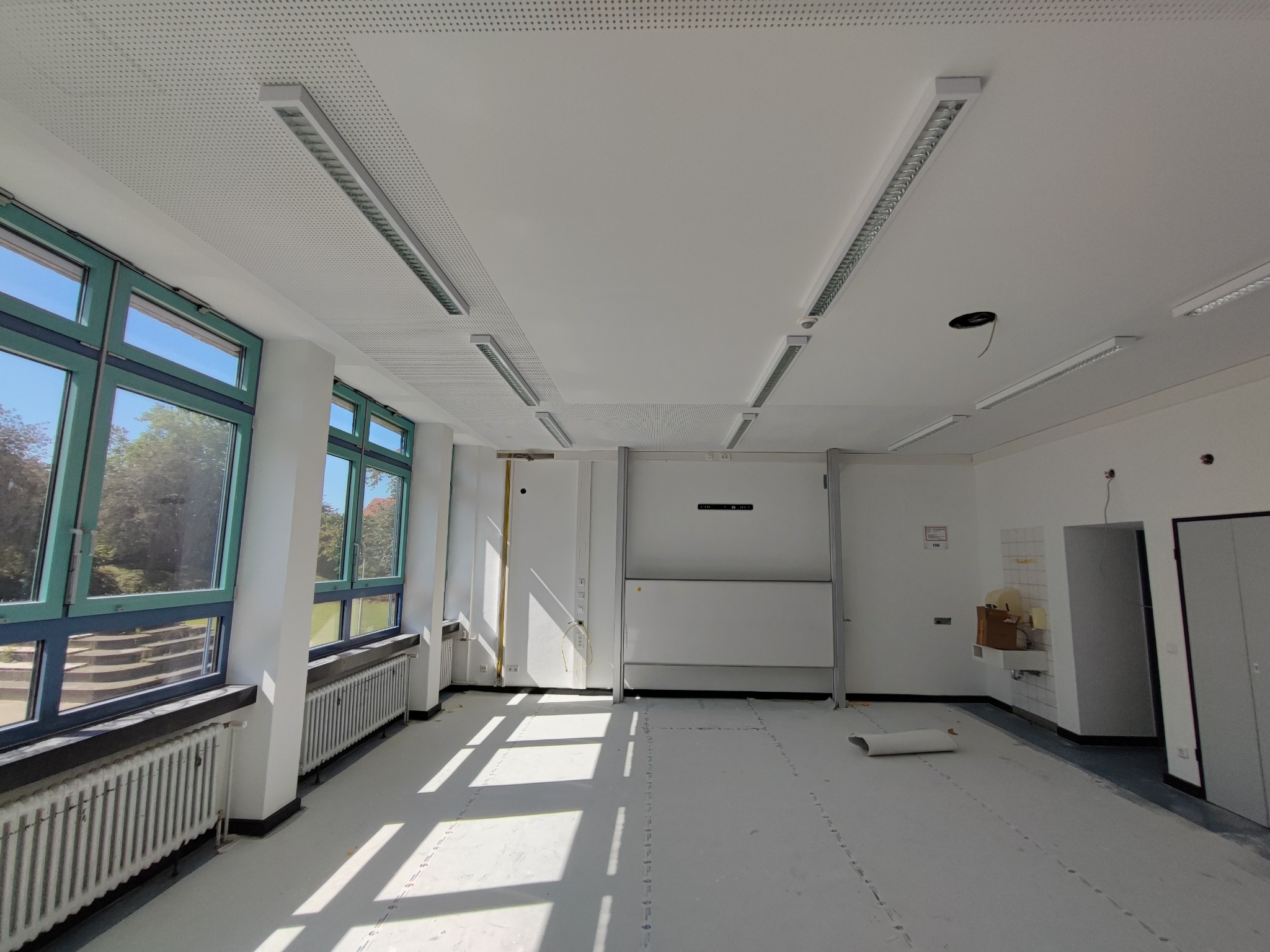 Przestronne, jasne pomieszczenie w trakcie prac wykończeniowych, widoczne okna z niebieskimi ramami, grzejniki pod oknami, biały sufit z oświetleniem liniowym i śladami po instalacji, przygotowane...