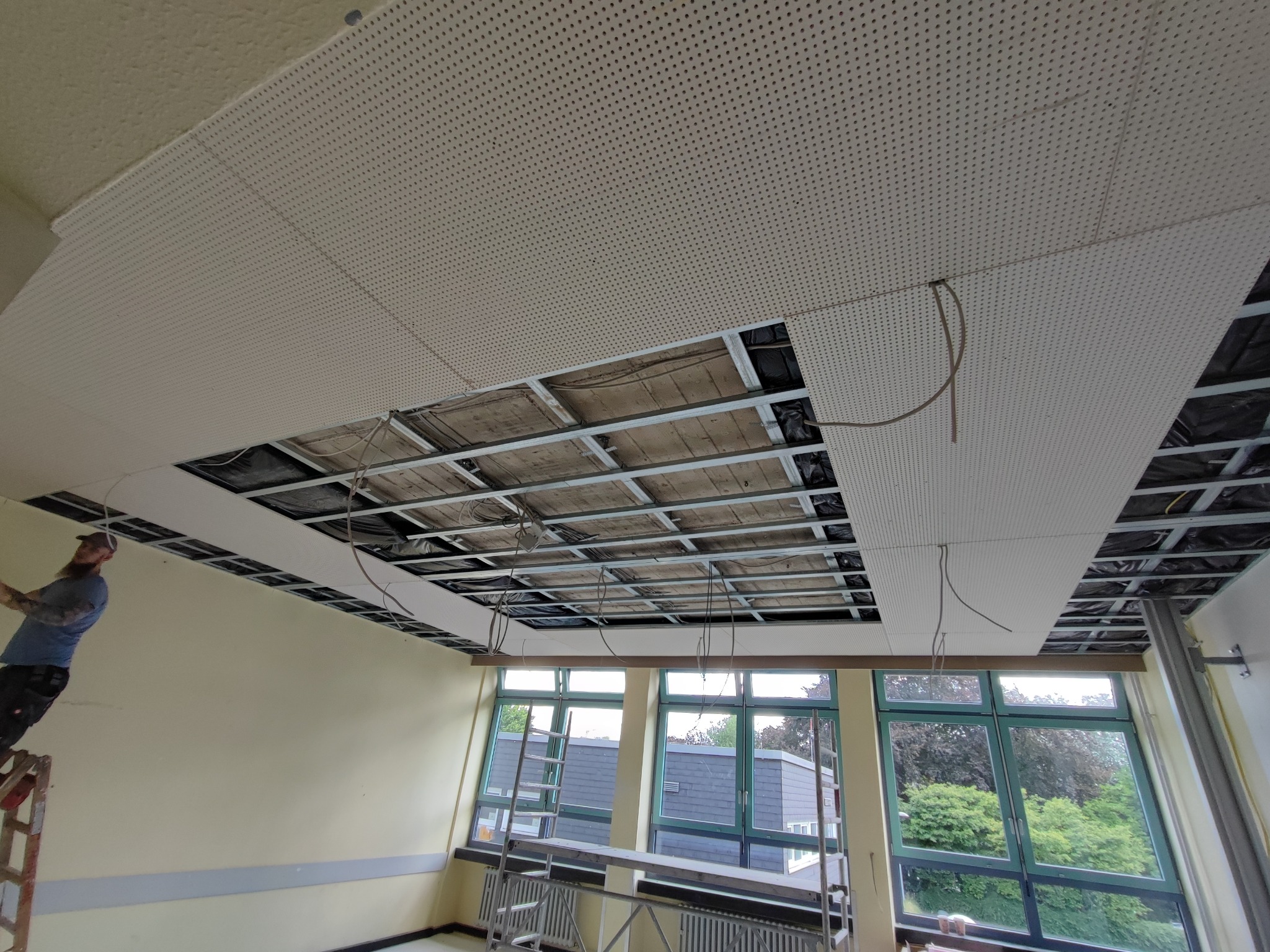 Montaż perforowanych paneli sufitowych na metalowej konstrukcji w pomieszczeniu z dużymi oknami; widoczny pracownik na drabinie oraz fragmenty instalacji elektrycznej.
