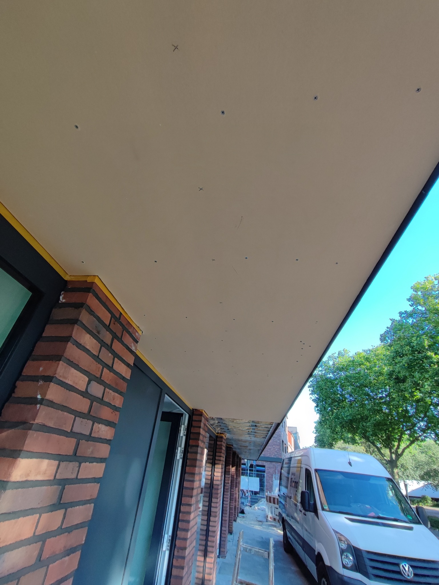 Niedokończony sufit podwieszany z płyt gipsowo-kartonowych, widoczne wkręty, ceglana elewacja budynku w tle, obok zaparkowany biały samochód dostawczy.