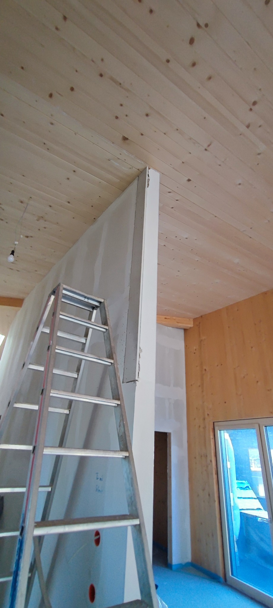 Wnętrze w trakcie prac wykończeniowych: ściana z płyt gipsowo-kartonowych, drewniany sufit, aluminiowa drabina oparta o ścianę, widoczne instalacje elektryczne w ścianie, fragment okna balkonowego...