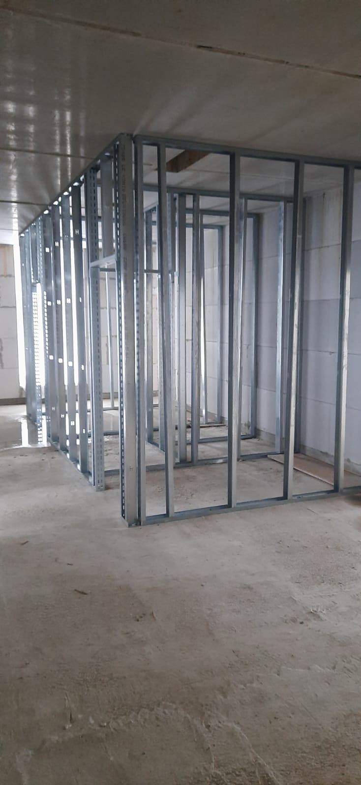 Metalowa konstrukcja szkieletowa ścian działowych w surowym stanie, widoczne profile stalowe, betonowa podłoga i bloczki betonowe w tle.