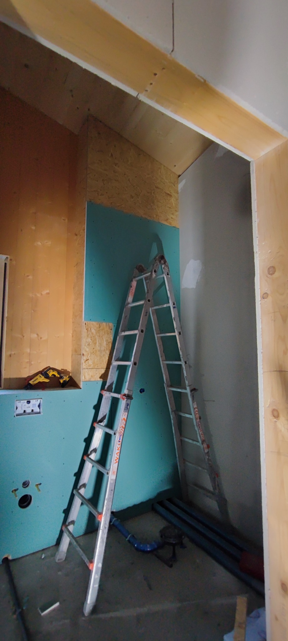 Wnętrze w trakcie prac wykończeniowych: widoczna drabina oparta o ścianę z zieloną płytą, obok instalacja hydrauliczna, narzędzia i fragment surowej drewnianej konstrukcji.
