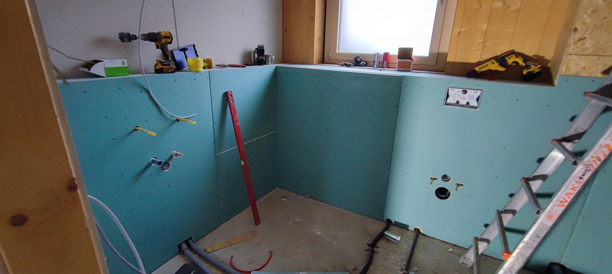 Wnętrze łazienki w Ostródzie w trakcie remontu, ściany wyłożone niebieską płytą, widoczne przygotowane punkty instalacji wodnej i odpływowej, poziomica oparta o ścianę, narzędzia na parapecie okna,...