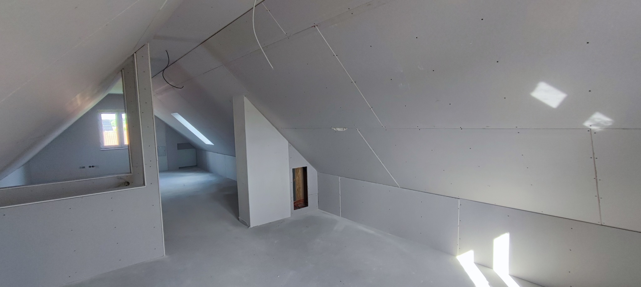 Poddasze po montażu płyt gipsowo-kartonowych, widoczne okno dachowe i otwór w ścianie, surowe wykończenie poddasza z widocznymi łączeniami płyt.