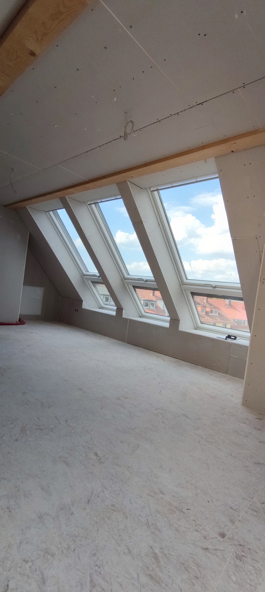 Poddasze z trzema oknami dachowymi, ściany i sufit wykończone płytami gipsowo-kartonowymi, widoczne łączenia płyt i drewniane elementy konstrukcyjne.