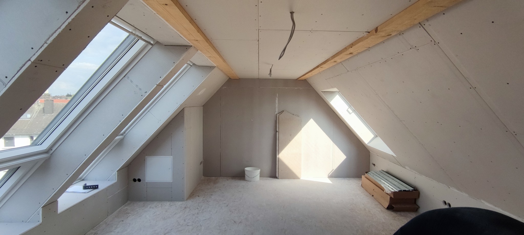 Poddasze w trakcie adaptacji: zamontowane płyty karton gips na skosach, drewniane belki stropowe, okna dachowe wpuszczające jasne światło, białe wiadro i profile metalowe na podłodze.