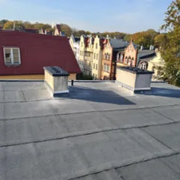 Widok z góry na płaski dach pokryty papą termozgrzewalną, z dwoma kominami wentylacyjnymi na tle zabudowy miejskiej i drzew w jesiennej kolorystyce.