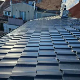 Nowy, błyszczący dach z ciemnoszarej blachodachówki modułowej na tle zabudowy miejskiej i błękitnego nieba z chmurami, widoczne fragmenty więźby dachowej.