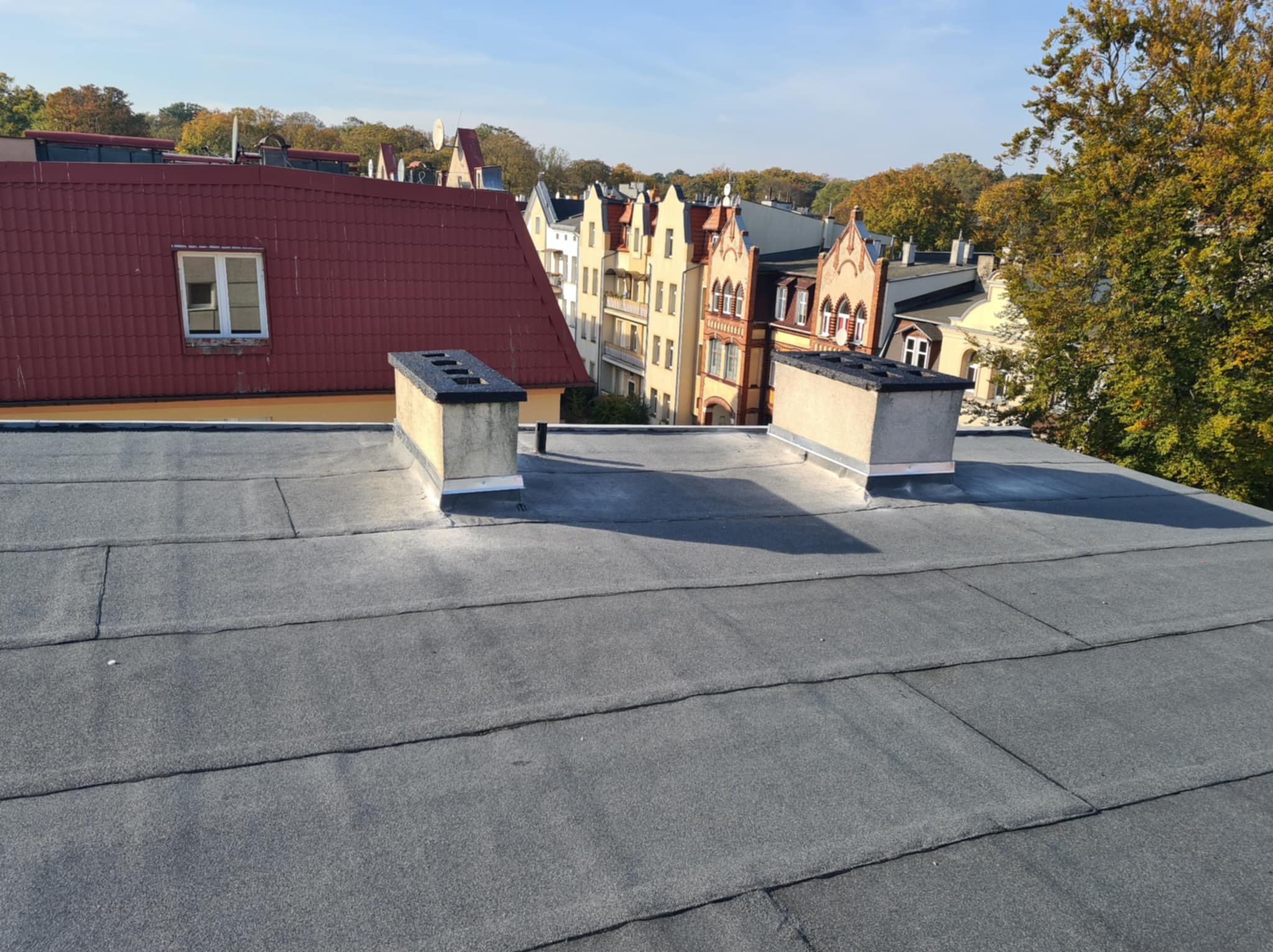 Widok z góry na płaski dach pokryty papą termozgrzewalną, z dwoma kominami wentylacyjnymi na tle zabudowy miejskiej i drzew w jesiennej kolorystyce.
