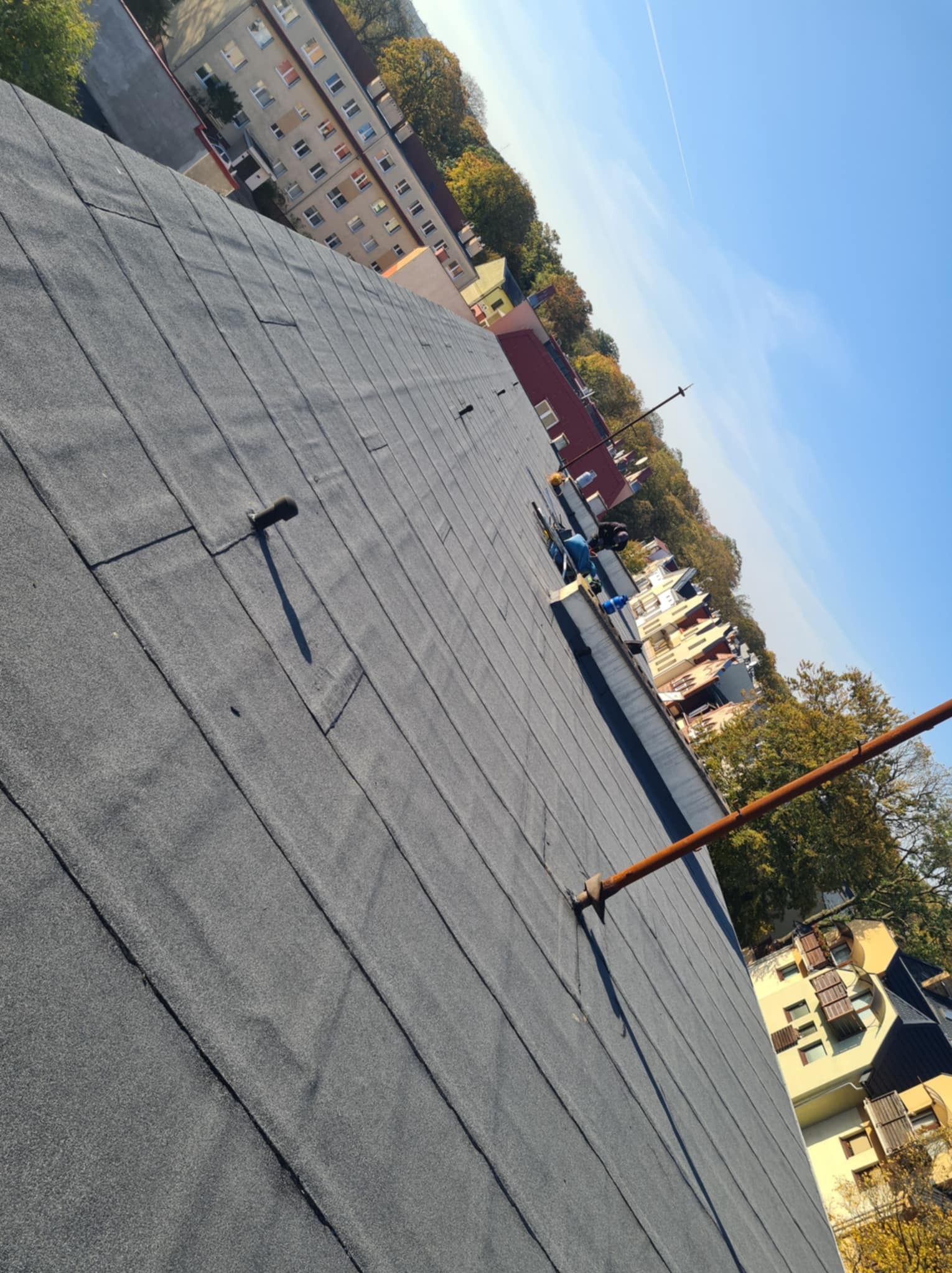 Dach pokryty papą termozgrzewalną na tle zabudowy miejskiej i błękitnego nieba, widoczny pion kanalizacyjny i pracownicy w oddali.