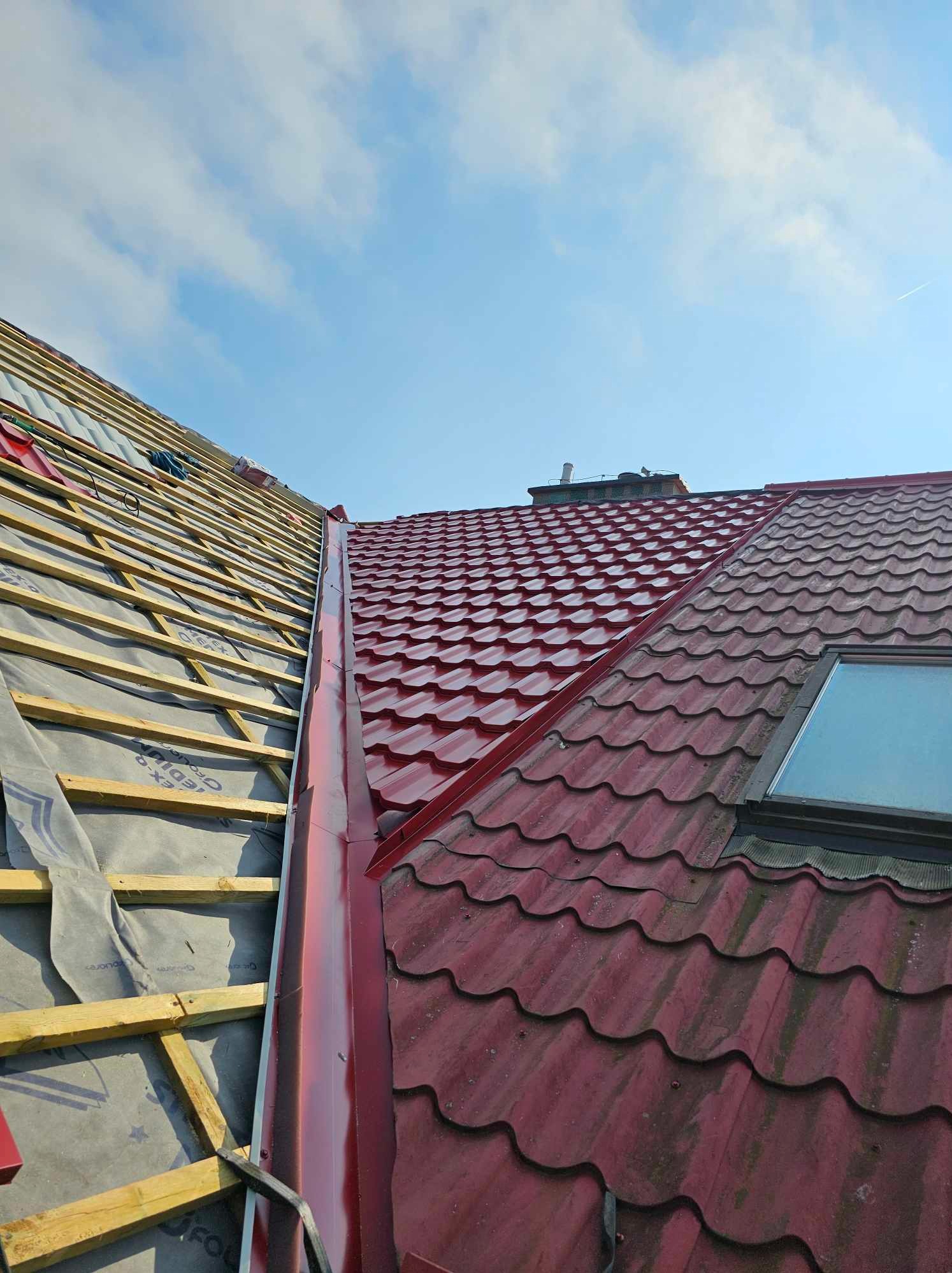 Częściowy widok dachu w Wolinie: połowa z drewnianą konstrukcją i membraną, druga połowa pokryta czerwoną blachodachówką z oknem dachowym i nową rynną.