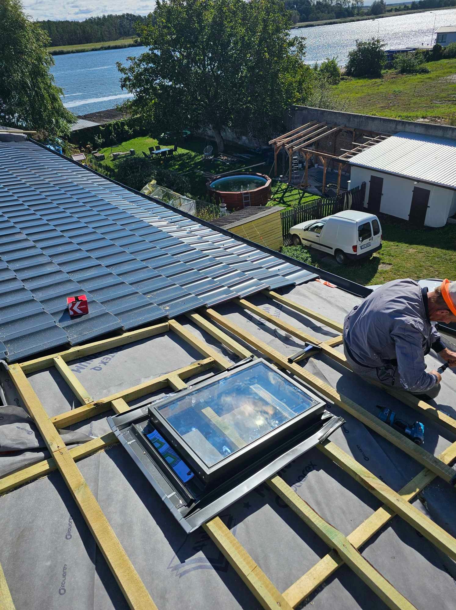Montaż okna dachowego na nowym dachu z szarą dachówką, widoczna więźba, membrana i narzędzia pracy dekarza, w tle jezioro i zabudowa.