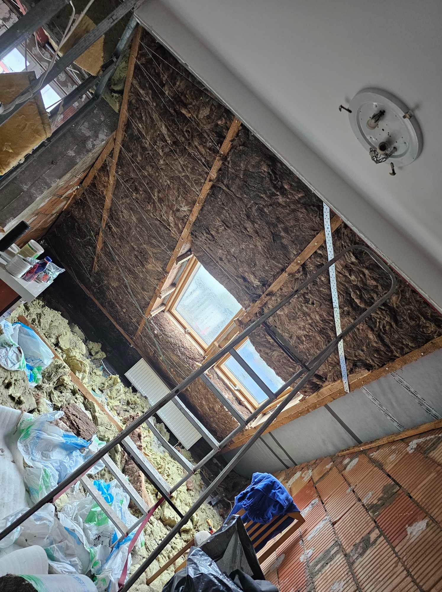 Poddasze w trakcie ocieplania, widoczne wełna mineralna, folia paroizolacyjna, drewniana konstrukcja, okno dachowe, drabina oraz worki z materiałem izolacyjnym.