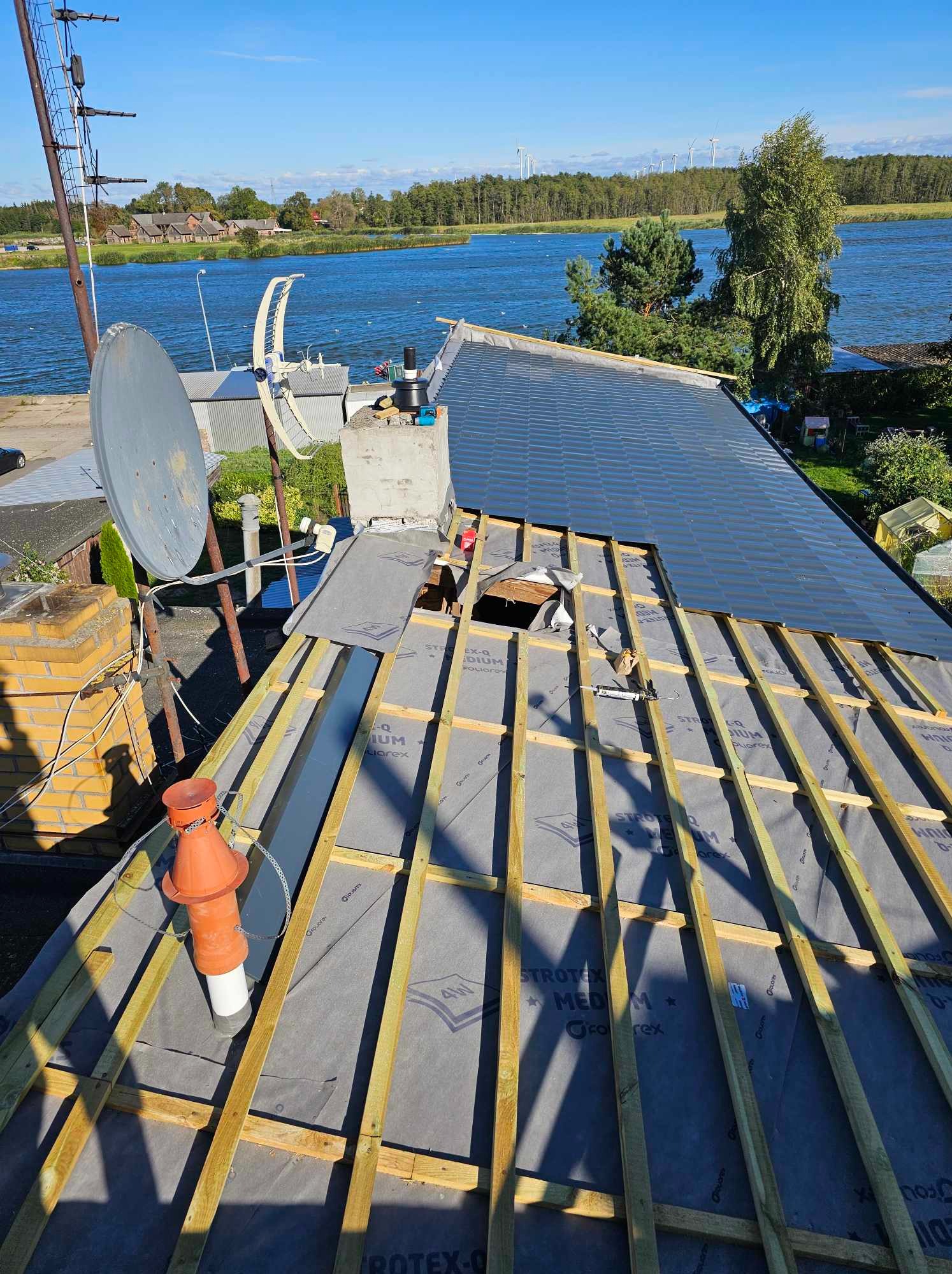 Dach z widoczną więźbą, membraną dachową i częściowo ułożonym pokryciem blachodachówką na tle błękitnego jeziora i turbin wiatrowych w oddali.
