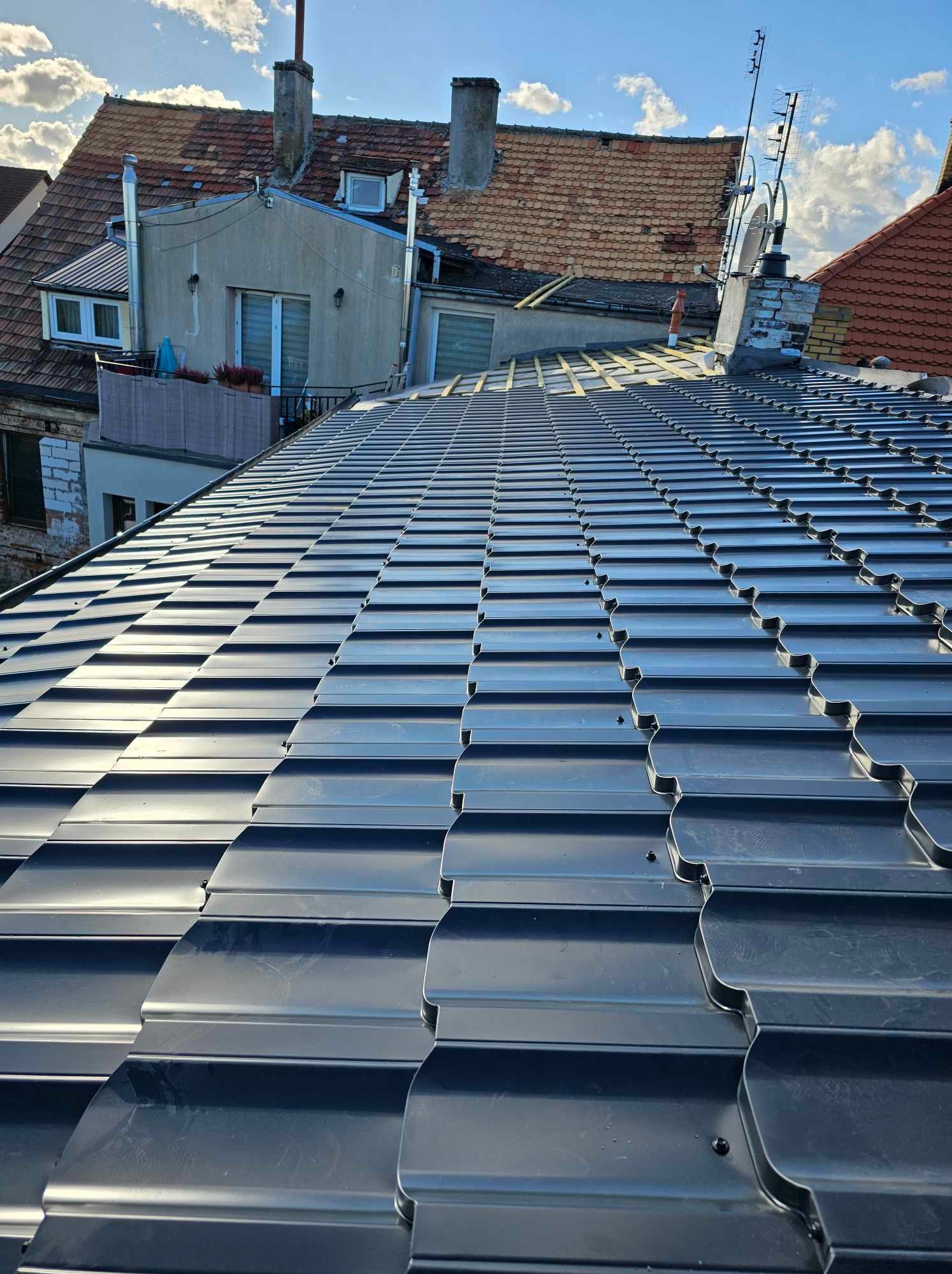 Nowy, błyszczący dach z ciemnoszarej blachodachówki modułowej na tle zabudowy miejskiej i błękitnego nieba z chmurami, widoczne fragmenty więźby dachowej.