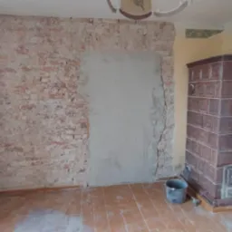 Ceglana ściana z fragmentem pokrytym świeżym tynkiem, brązowy piec kaflowy w rogu pokoju, widoczne ślady remontu na drewnianej podłodze i wiadro na podłodze.