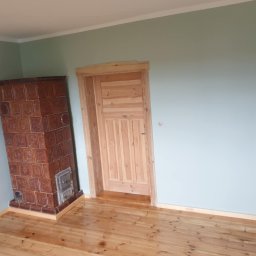 MattBud - Wnętrze pokoju po remoncie: świeżo pomalowane ściany w odcieniach szarości, nowa drewniana podłoga, piec kaflowy z brązowych płytek oraz surowe drewniane drzwi z ościeżnicą.