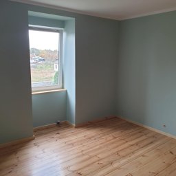 MattBud - Pomalowane na pastelowo zielono wnętrze pokoju z drewnianą podłogą, widok na zewnątrz przez okno z drewnianym parapetem, w rogu pod oknem stoi puszka z farbą.